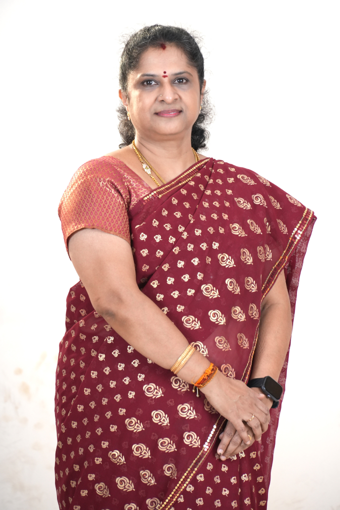 Portrait of a woman with dark curly hair tied back, wearing a bright orange and green silk saree with floral patterns. She is wearing gold jewelry, including necklaces and earrings, and has a red bindi on her forehead. The photograph is a formal headshot against a plain light-colored background.