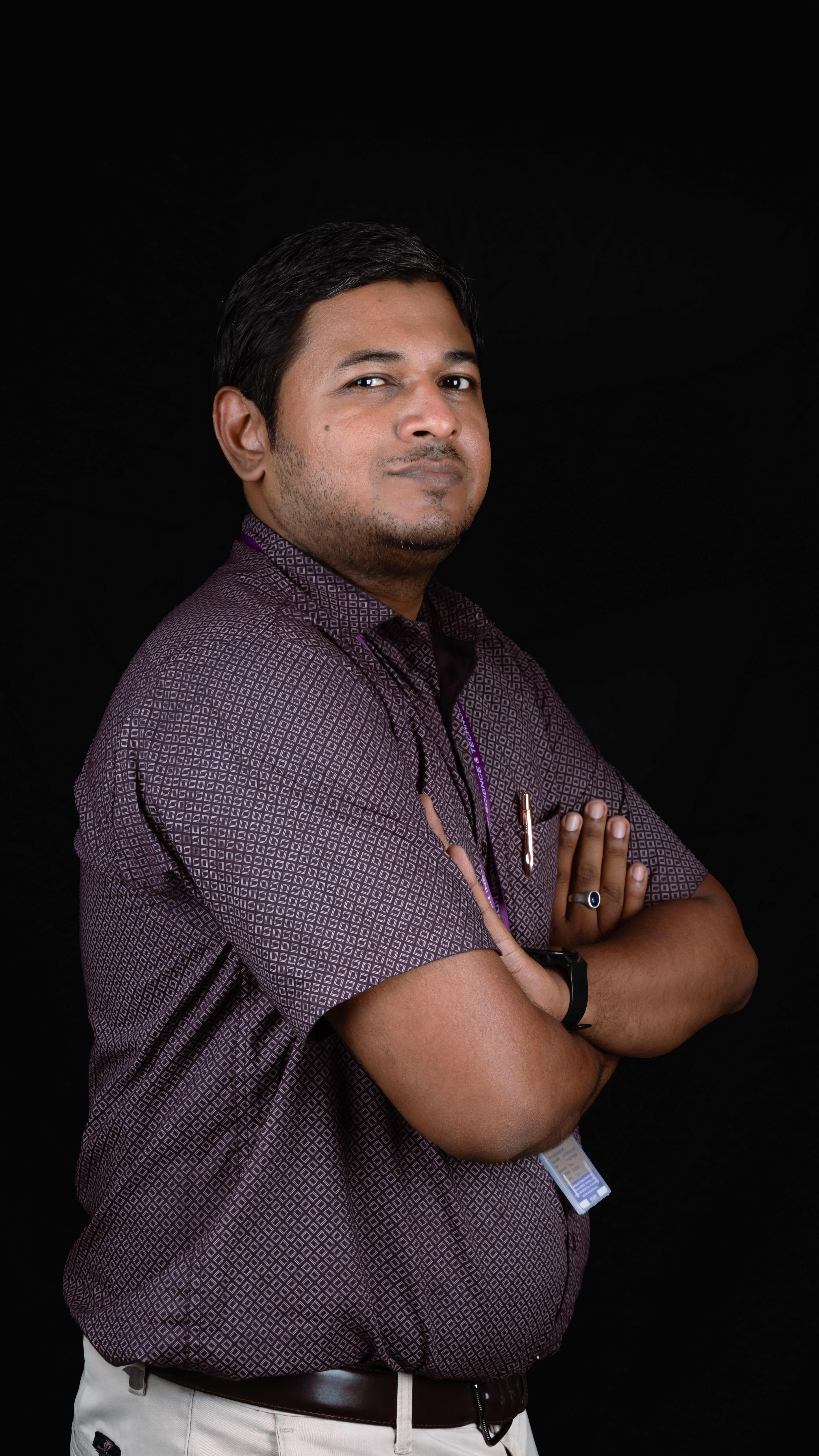 Studio portrait of a man standing with his arms crossed against a black background. He is wearing a patterned dark maroon short-sleeved button-down shirt tucked into light-colored trousers with a dark brown belt. A staff ID card is partially visible on a lanyard around his neck. He has short black hair, light facial hair, and a calm, confident expression while looking slightly toward the camera. He is also wearing a wristwatch and a ring.