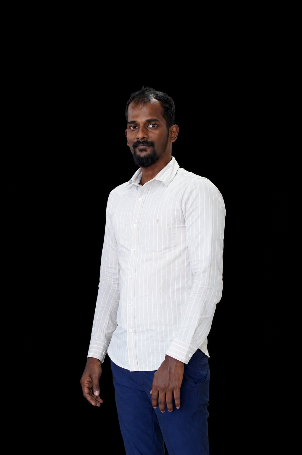 Studio portrait of a man standing against a black background. He is wearing a white long-sleeved button-down shirt with subtle vertical stripes and dark blue trousers. He has short black hair, a mustache, and a pointed goatee beard. His arms rest naturally at his sides, and he faces slightly toward the camera with a calm, confident expression