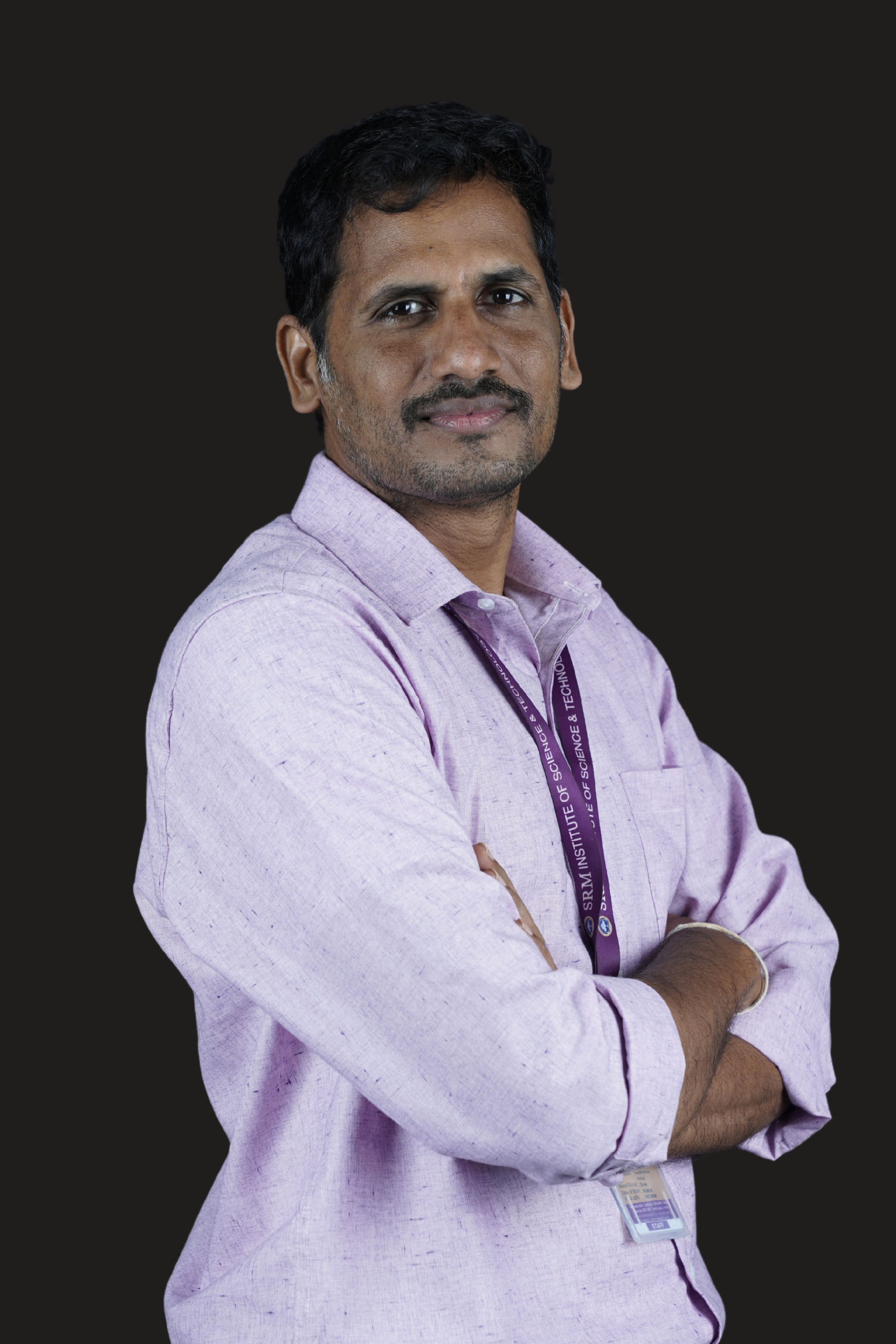 Studio portrait of a man standing with his arms crossed against a dark background. He is wearing a light lavender long-sleeved button-down shirt and a purple lanyard with a staff ID card. He has short black hair, a mustache with light facial stubble, and a slight smile while looking directly at the camera. The pose conveys confidence and professionalism.