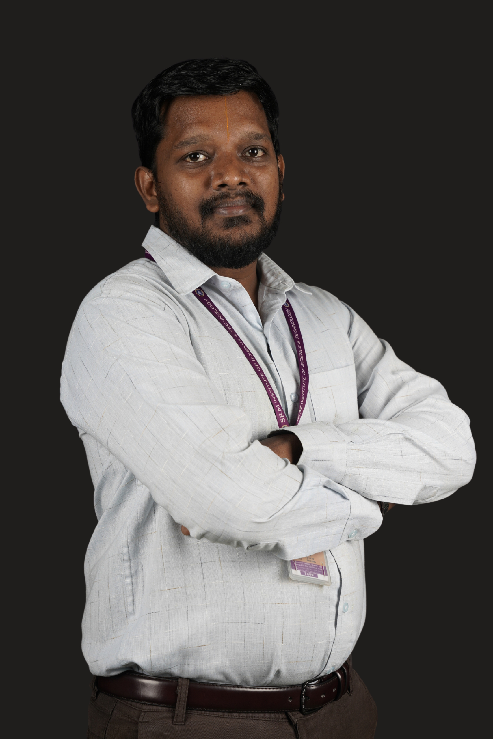Studio portrait of a man standing with his arms crossed against a dark background. He is wearing a light gray long-sleeved button-down shirt tucked into dark trousers with a brown belt. A staff ID card on a purple lanyard hangs around his neck. He has short black hair, a beard and mustache, and a calm, confident expression while looking toward the camera.