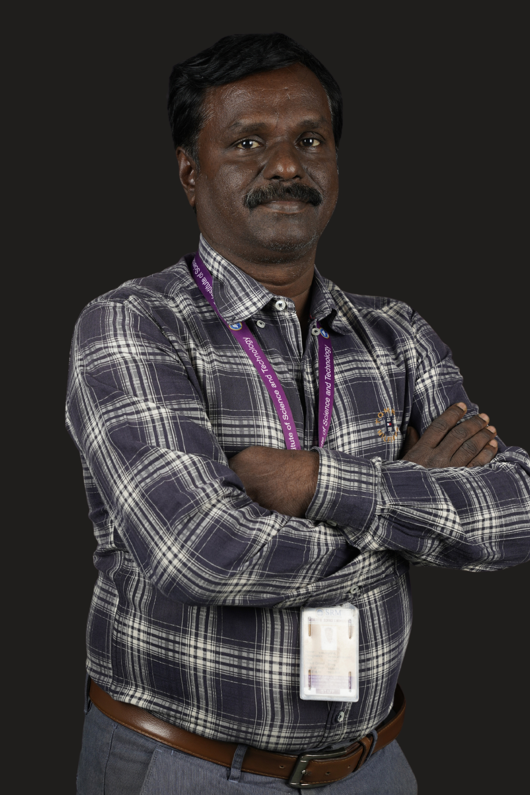 Studio portrait of a man standing with his arms crossed against a dark background. He is wearing a dark blue and white plaid long-sleeved button-down shirt tucked into gray trousers with a brown belt. A staff ID card on a purple lanyard hangs around his neck. He has short black hair, a mustache, and a calm, professional expression while looking directly at the camera. The pose conveys confidence and approachability.